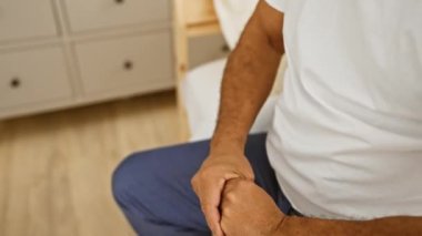 Yatakta oturan orta yaşlı bir adamın yakın plan fotoğrafı. Elleri sıcak ışıklandırmalı bir yatak odasına sıkışmış..