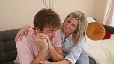 Anne ve oğul destekleyici bir kucaklaşmayı paylaşıyor, evdeki kanepede oturuyor, aile üzüntü ve endişeyle başa çıkıyor.