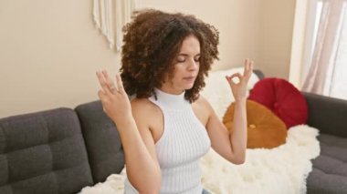 Kıvırcık saçlı, ışıl ışıl Latin bir kadın rahatına sarılıyor evdeki kanepesinde rahatça oturuyor. Gözleri huzurlu bir şekilde kapalıyken, rahatlatıcı bir yoga meditasyon hareketi yapıyor..
