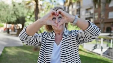 Mutlu, olgun sarışın kadın şehir parkında elleriyle kalp işareti yapıyor.