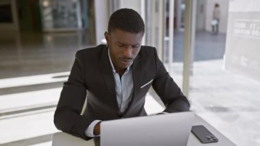Modern ofis ortamında dizüstü bilgisayar kullanan odaklanmış siyahi adam, profesyonellik ve konsantrasyon tasvir ediyor.