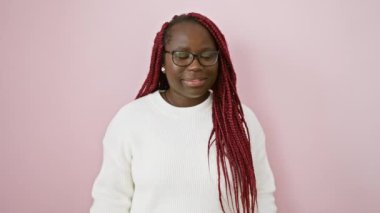 Örgülü ve gözlüklü neşeli bir Afrikalı kadın öne bakıyor, pembe arka planda beyaz bir kazak giyiyor..