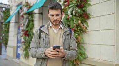 Sakallı genç bir İspanyol, dekore edilmiş bir caddede telefonunu kontrol ediyor..