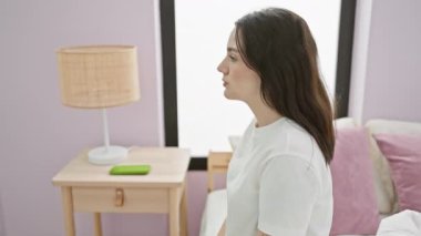 Pijamalı, kendine güvenen, doğal bir şekilde gülümseyen, yatakta oturmuş, yatak odasına bakan, kendine güvenen genç bir kadın..