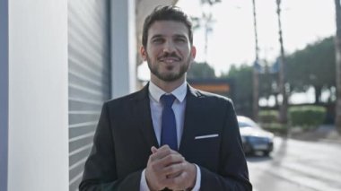 Gülümseyen profesyonel takım elbiseli adam güneşli bir caddede duruyor arkasında binalar ve araba var.