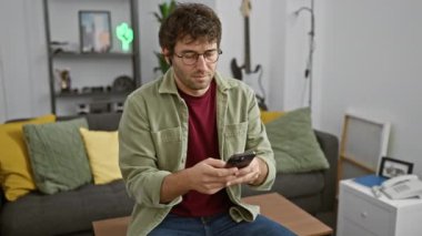 Kulaklık ve gözlüklü genç bir İspanyol evde akıllı telefon kullanıyor, rahat ve odaklanmış bir atmosfer yayıyor..