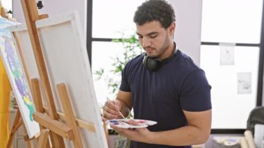 Parlak bir sanat stüdyosunun tuvaline sakallı, kulaklık takan dalgın bir adam.