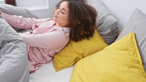 カーリーヘアの若い女性は ピンクのナイトガウンと黄色の枕で彼女の寝室に伸びる — ストック動画