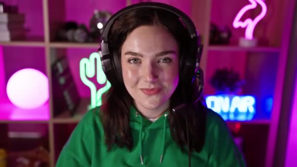 一位身穿绿色帽衫 头戴耳机 面带微笑的年轻女子 晚上在霓虹灯下的游戏室里 — 图库视频影像