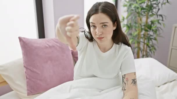 怒っている若い女性がベッドに座って 彼女のパジャマで親指のジェスチャーに嫌悪感を示し 屋内の気分の悪い肖像画 — ストック動画