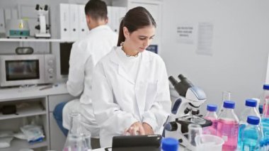 Laboratuvarda bir kadın ve bir erkek, bilimsel ekipman ve cam parçalarıyla çevrili örnekleri birlikte analiz ediyor..