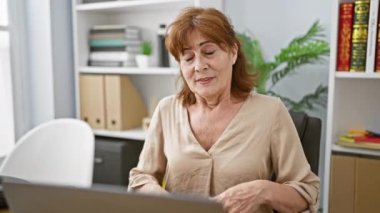 Mutlu orta yaşlı bir kadın, minnettar hissediyor, sağlık dolu içten gülümsüyor, ellerini göğsüne koyuyor, koltukta otururken, dizüstü bilgisayarını kullanırken minnettarlık göstergesi olarak..