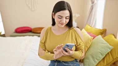 Hispanic woman smiles while texting on a smartphone in a cozy, well-lit bedroom with colorful pillows.