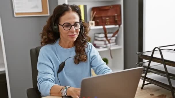 ヘッドフォンを着用しているカーリーヘアの笑顔のヒスパニック女性は 現代のオフィスのインテリアで彼女のラップトップで動作します — ストック動画