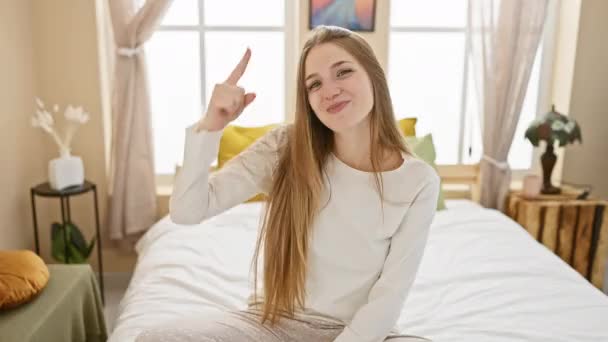 パジャマの美しい若い金髪の女性 幸せにベッドに座って 喜んで彼女のヘッドウィンキングを指す 彼女の居心地の良い寝室で素晴らしいアイデアや良い記憶 — ストック動画