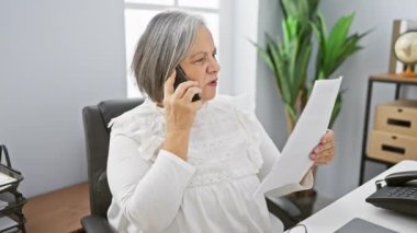 Üst düzey bir kadın modern bir ofiste telefonla konuşurken bir belgeyi inceliyor..