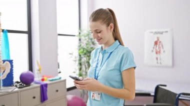 Üniformalı, gülümseyen kadın terapist. Parlak bir rehabilitasyon merkezinde tıbbi ekipmanla akıllı telefon kullanıyor.