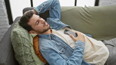 Kapalı bir koltukta uzanırken akıllı telefon kullanan sakallı, rahat, genç bir İspanyol..