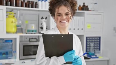 Kıvırcık saçlı, laboratuvar önlüğü ve eldiveni giyen neşeli bir İspanyol kadın laboratuvar ortamında pano tutuyor..