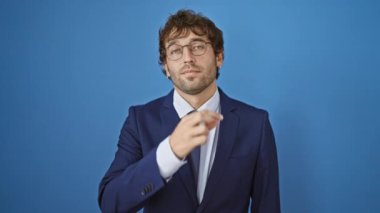 Genç adamın sırrı, iş elbisesi, parmaklarının fermuarı kapalı. Mavi izole arka planda sessiz bir jest. Sessiz iletişim, ciddi ifade, gizli konsept