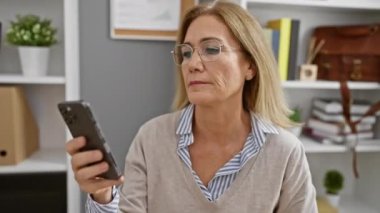 Modern ofis içinde akıllı telefon kullanan, gözlüklü olgun bir iş kadını endişe ve memnuniyeti dile getiriyor..