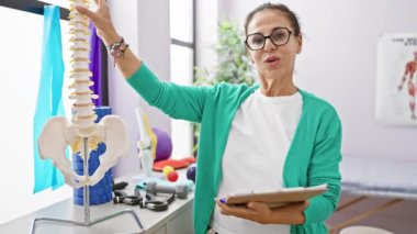 Gülümseyen, olgun, gözlüklü bir İspanyol kadın terapist, parlak bir rehabilitasyon kliniğinde omurga modeli gösteriyor..