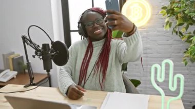 Örgülü Afrikalı Amerikalı bir kadın stüdyoda mikrofon ve neon ışıklarla selfie çekiyor..