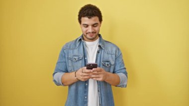 Heyecanlı genç Arap adam elinde akıllı telefonuyla sevinçle işaret ederek izole edilmiş sarı arka plan hakkında olumlu bir fikir gösteriyor. Sakalları ışıl ışıl, iyimser ve kendine güvenen neşeli Arap erkek..