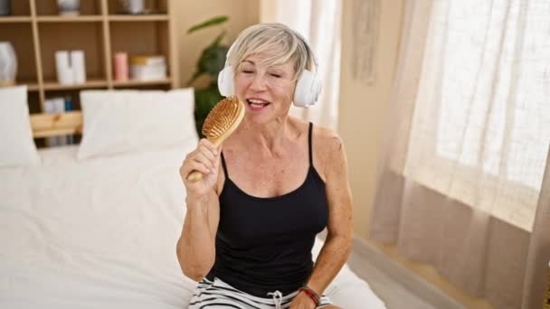 拿着耳机的成熟女人一边拿着梳子一边在卧室的床上唱歌 一边欣赏着音乐 — 图库视频影像