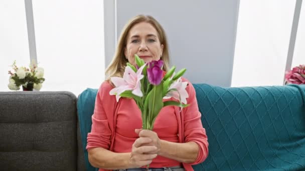 居心地の良いリビングルームでピンクと紫の花の花束を保持する笑顔の中年の女性 — ストック動画