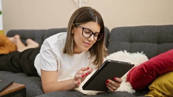 一位带着眼镜的惊慌失措的年轻女人躺在家里的沙发上 一边笑一边看平板电脑 — 图库视频影像
