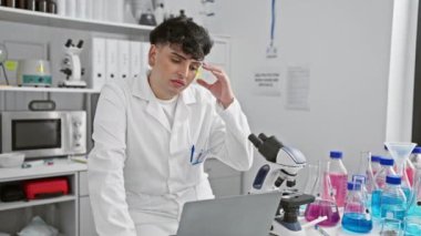 Laboratuvar ortamındaki genç bir adam, mikroskop ve renkli cam parçalarının yanındaki dizüstü bilgisayarda çalışırken stresli görünüyor..