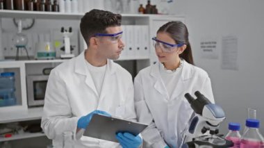 Laboratuvar önlüğü ve güvenlik gözlüğü takan bir kadın ve erkek bilim adamı laboratuvar ortamında ekipmanlarla bir tablet üzerinde tartışıyor..