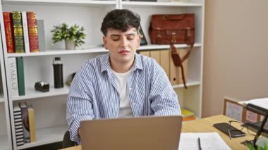 Çizgili tişörtlü genç bir adam, modern ofis ortamında dizüstü bilgisayarının başında çalışırken yorgunluğunu ifade ediyor..