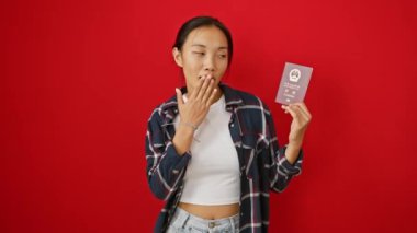 Şok olmuş genç Çinli kadın ağzını korkuyla kapatıyor, Çin pasaportu tutuyor, kırmızı arka planda şaşırmış bir ifade sergiliyor.