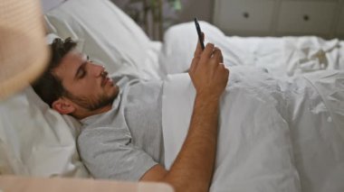 Yatakta sakallı, evde akıllı telefon kullanan, yatak odası düzeninde boş vaktini ve teknolojisini sergileyen Latin kökenli genç bir adam.