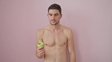 Tişörtsüz İspanyol genç adam, gülümseyen, mutluluğun tadını çıkaran, çapraz kollu bir elma tutan, serin pembe bir arka plan üzerinde sağlık ve pozitiflik bir resim.