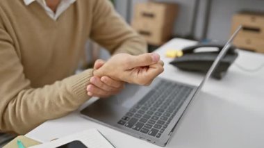 Ofis ortamında sakallı, dizüstü bilgisayar kullanırken bileği ağrıyan genç bir İspanyol..