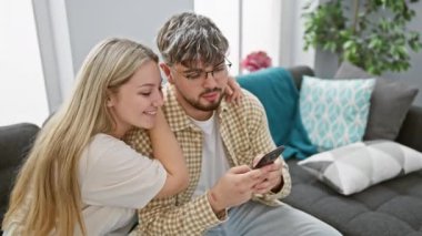 Bir çift evdeki kanepede rahat bir anın tadını çıkarıyor. Erkek, nişanlı kadına akıllı telefondan bir şey gösteriyor..