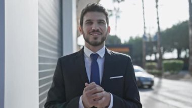 Gülümseyen profesyonel takım elbiseli adam güneşli bir caddede duruyor arkasında binalar ve araba var.