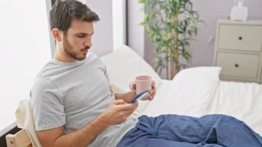 Yakışıklı İspanyol bir adam kahve içiyor ve yatak odasında akıllı telefon okuyor.