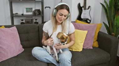 Kulaklıklı rahat bir kadın renkli yastıklarla çevrili rahat bir koltukta otururken köpeğini nazikçe tutar..