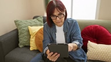 Olgun İspanyol kadın evdeki kanepede oturuyor, odaklanmış ve rahat bir tablet kullanıyor.