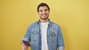 Neşeli, genç, Arap bir adam, elinde emirlik pasaportu, gülümsüyor, sarı izole edilmiş arka planda mutlu, dişlek bir gülümseme