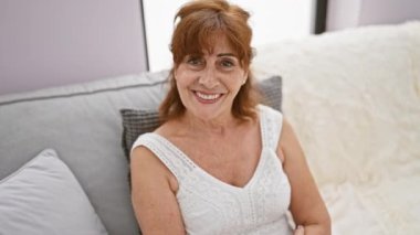 Neşeli orta yaşlı kadın, kendine güvenen kızıl saçlı, olumlu duygularını ifade eden, başparmağını kaldırmış, kanepeye oturmuş elbise giymiş, evdeki rahat oturma odasında ağzı açık bir şekilde ışıldıyor.