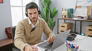 Kulaklık takan odaklı İspanyol bir adam modern ofis ortamında dizüstü bilgisayarında çalışır..
