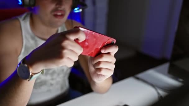 一个专心致志的年轻人晚上在黑暗的房间里玩耍 戴着耳机 手持红色的智能手机 — 图库视频影像