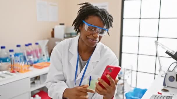 快乐的年轻黑人女人 头发蓬乱 在实验室里工作 自信地使用智能手机 开心地微笑着指着自己 — 图库视频影像