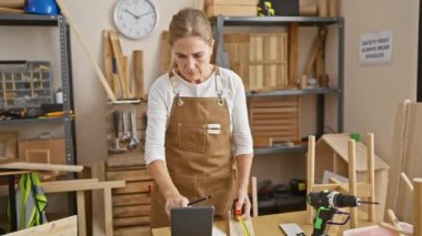 Marangozluk atölyesinde elinde aletlerle tablet kullanan olgun sarışın bir kadın..