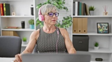 Gri saçlı olgun bir kadın organize bir ofiste pembe kulaklıklarla müzik dinliyor..